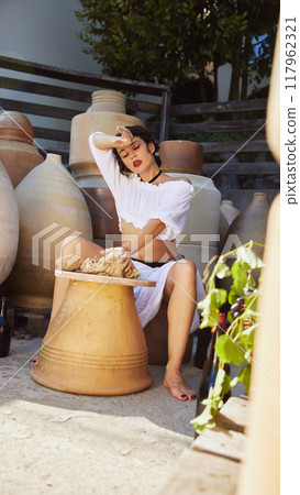 Elegant young woman in traditional clothing, shaping clay with focused expression in sunlit pottery studio, large jars around her. Concept of travelling. Elegant young woman in traditional clothing, shaping clay with focused expression in sunlit pottery studio, large jars around her. Concept of travelling. 117962321