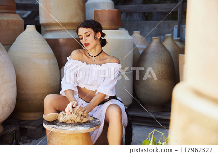 Young, beautiful woman with dark hair in historical attire, working clay in pottery workshop surrounded by large earthenware jars. Concept of travelling. Young, beautiful woman with dark hair in historical attire, working clay in pottery workshop surrounded by large earthenware jars. Concept of travelling. 117962322