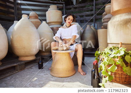 Young woman in white dress, works with clay surrounded by pottery, blending artistry of ceramics with rustic beauty of Mediterranean tradition. Concept of travelling. 117962323
