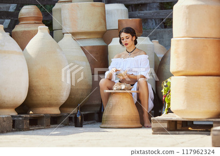 Young woman in white dress, molds clay among large terracotta pots, embracing artisanal spirit and cultural history of Mediterranean pottery. Concept of travelling. 117962324
