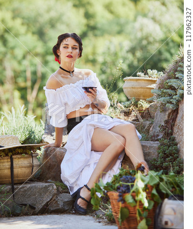 Young woman in white dress, rests with glass of wine, her surroundings celebration of Mediterranean harvest and beauty of countryside. Concept of travelling. 117962327