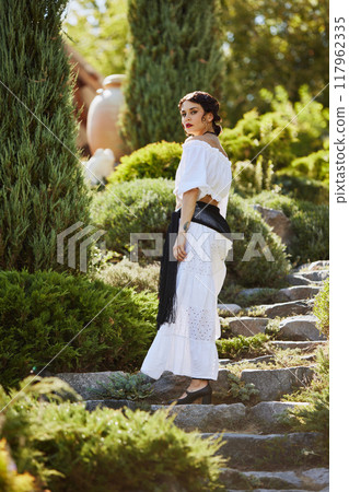 Young woman with dark hair in white dress, turns on stone steps, surrounded by lush greenery. Timeless beauty of Mediterranean garden. Concept of harvesting. 117962335