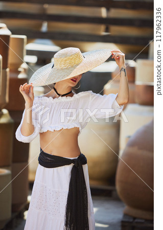 Young woman adjusts her hat, preparing to face autumn sun, her surroundings steeped in rich history of Mediterranean pottery art. Concept of harvesting. Young woman adjusts her hat, preparing to face autumn sun, her surroundings steeped in rich history of Mediterranean pottery art. Concept of harvesting. 117962336