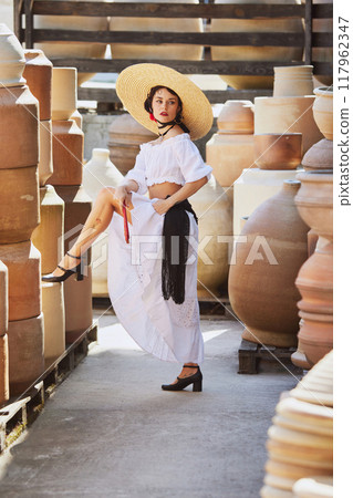 Young woman with dark hair, in white dress, poses gracefully among large terracotta pots in Spanish pottery yard, celebrating autumn's artisanal beauty. 117962347