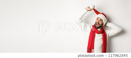 Winter holidays and New Year party concept. Happy bearded man celebrating Christmas, playing with Santa hat and looking up, white background 117962845