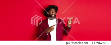Winter holidays and shopping concept. Happy Black man pointing left and smiling, showing new year promo offer on red background 117962850
