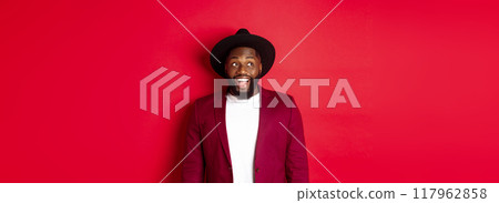 Fashion and party concept. Excited african american man looking at logo, staring upper left corner with happy smile, standing against red background 117962858
