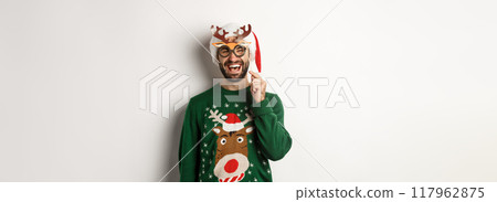 Christmas and holidays concept. Smiling beared man in Santa hat looking happy, holding party mask for New Year, celebrating xmas, standing over white background 117962875