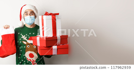 Winter holidays and xmas concept. Happy man in face mask and santa hat bringing gifts, holding Christmas sock and present boxes, standing over white background Winter holidays and xmas concept. Happy man in face mask and santa hat bringing gifts, holding Christmas sock and present boxes, standing over white background 117962939