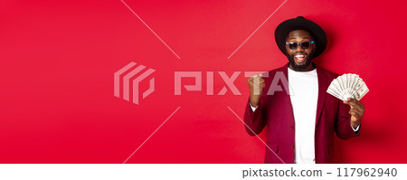 Cheerful african american guy in sunglasses, black hat and blazer, winning prize money, holding dollars and looking satisfied, standing over red background Cheerful african american guy in sunglasses, black hat and blazer, winning prize money, holding dollars and looking satisfied, standing over red background 117962940