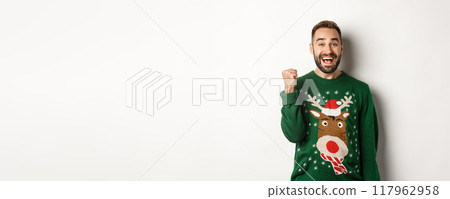 New Year, holidays and celebration. Excited and happy guy in Christmas sweater making fist pump, triumphing, receiving a gift and rejoicing, standing over white background New Year, holidays and celebration. Excited and happy guy in Christmas sweater making fist pump, triumphing, receiving a gift and rejoicing, standing over white background 117962958