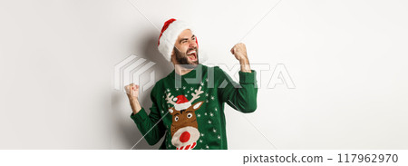 Christmas holidays, celebration and party concept. Happy guy in Santa hat and sweater, making fist pumps and rejoicing, triumphing, standing over white background 117962970