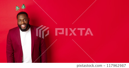 Merry Christmas. Handsome african american man in blazer and party headband, celebrating new year, smiling happy at camera, red background 117962987