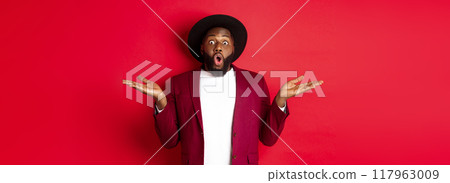 Christmas shopping and people concept. Handsome african american man staring amazed, spread hands sideways, showing promo offers on copy space, red background 117963009