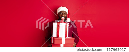 Christmas, New Year and shopping concept. Happy Black man in santa hat and blazer holding xmas presents, bring gifts and smiling, standing against red background 117963017