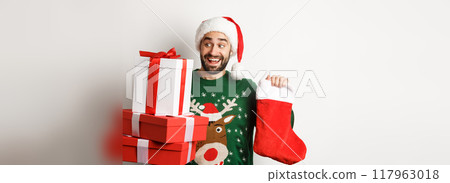 Xmas and winter holidays concept. Excited man holding christmas sock and gift boxes, celebrating New Year, standing over white background 117963018