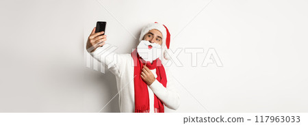 Christmas party and celebration concept. Young man taking selfie with funny white beard Santa mask and hat, posing for photo on mobile phone, studio background 117963033