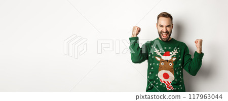 New Year, holidays and celebration. Excited bearded man in Christmas sweater, making fist pumps and shouting for joy, rejoicing and triumphing, white background New Year, holidays and celebration. Excited bearded man in Christmas sweater, making fist pumps and shouting for joy, rejoicing and triumphing, white background 117963044