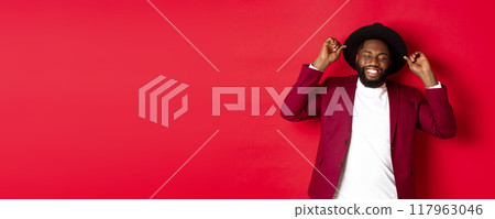 Christmas shopping and people concept. Happy Black man celebrating new year, wearing party outfit, touching hat and smiling, standing against red background Christmas shopping and people concept. Happy Black man celebrating new year, wearing party outfit, touching hat and smiling, standing against red background 117963046