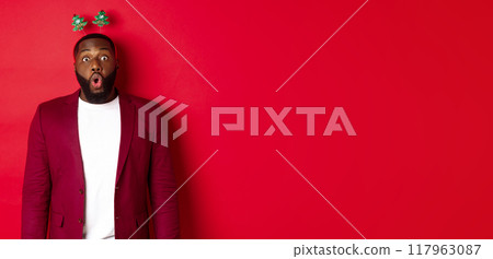 Merry Christmas. Happy african american man celebarting New Year, wearing party headband and making funny faces , standing over red background Merry Christmas. Happy african american man celebarting New Year, wearing party headband and making funny faces , standing over red background 117963087