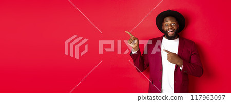 Winter holidays and shopping concept. Happy Black man pointing left and smiling, showing new year promo offer on red background Winter holidays and shopping concept. Happy Black man pointing left and smiling, showing new year promo offer on red background 117963097
