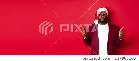 Funny Black man celebrating New Year, wearing party glasses and santa hat, showing thumb up, approve and like, standing over red background 117963100