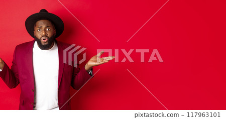 Confused and worried Black man spread hands sideways, staring at camera puzzled, asking what wrong, standing against red background Confused and worried Black man spread hands sideways, staring at camera puzzled, asking what wrong, standing against red background 117963101