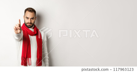 Serious man saying no, showing stop sign, shaking finger in decline, rejection sign, prohibit action, standing in winter sweater and red scarf against white background Serious man saying no, showing stop sign, shaking finger in decline, rejection sign, prohibit action, standing in winter sweater and red scarf against white background 117963123