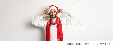 Winter holidays and New Year party concept. Happy man celebrating Christmas, wearing santa hat and red scarf, laughing out loud, white background 117963127