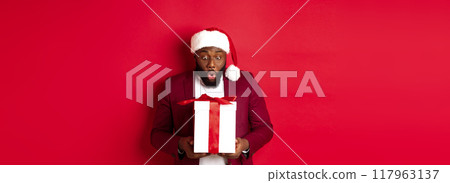 Christmas, New Year and shopping concept. Surprised Black man staring at xmas present, saying wow amazed, receiving holiday gift, red background 117963137