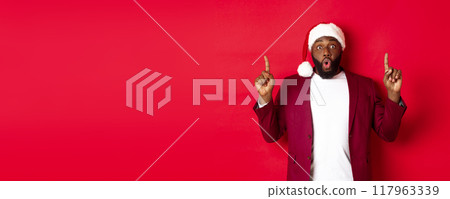 Christmas, party and holidays concept. Impressed Black man showing offer, pointing fingers up at logo, saying wow, standing against red background Christmas, party and holidays concept. Impressed Black man showing offer, pointing fingers up at logo, saying wow, standing against red background 117963339