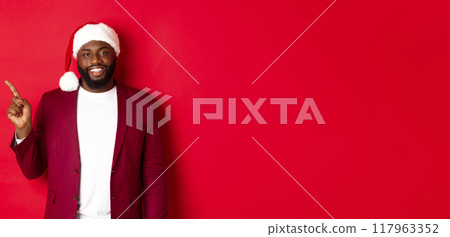 Christmas, party and holidays concept. Handsome Black man in santa hat pointing finger left, showing advertisement, standing happy over red background 117963352
