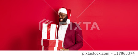 Christmas, New Year and shopping concept. Happy african amaerican man looking touched and thankful at xmas presents, receiving gifts, wearing santa hat, red background 117963374