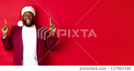 Christmas, party and holidays concept. Excited Black man in santa hat and blazer, pointing fingers up and smiling, showing promo offer, red background 117963380