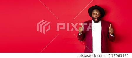 Christmas shopping and people concept. Handsome Black man smiling satisfied, showing thumbs-up, like and agree, approve something, standing against red background 117963381