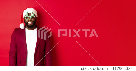 Funny Black man in santa hat and party glasses celebrating Christmas, smiling happy and wishing merry xmas, standing over red background Funny Black man in santa hat and party glasses celebrating Christmas, smiling happy and wishing merry xmas, standing over red background 117963385