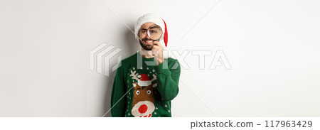 Christmas and holidays concept. Happy bearded man in Santa hat and xmas sweater, looking through magnifying glass and smiling, standing over white background 117963429