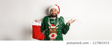 Xmas and winter holidays concept. Confused guy in Santa hat holding Christmas sock and shrugging, standing clueless against white background 117963433