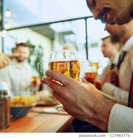Focus on male hand holding lager beer. Business people, colleagues meeting at local bar, drinking beer, talking and celebrating successful collaboration Focus on male hand holding lager beer. Business people, colleagues meeting at local bar, drinking beer, talking and celebrating successful collaboration 117963778