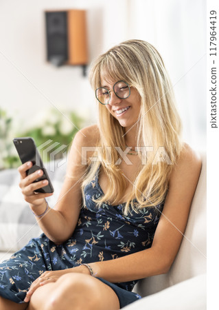 Happy relaxed young woman sitting on couch using cell phone, smiling lady laughing holding smartphone, looking at cellphone enjoying doing online ecommerce shopping in mobile apps or watching videos. Happy relaxed young woman sitting on couch using cell phone, smiling lady laughing holding smartphone, looking at cellphone enjoying doing online ecommerce shopping in mobile apps or watching videos. 117964419