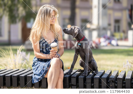 A young woman sits in a city park with her dog, a Weimaraner A young woman sits in a city park with her dog, a Weimaraner 117964430