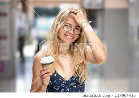 A good-looking blonde in a summer outfit with glasses is walking through the city, holding a take away coffee in her hand 117964436