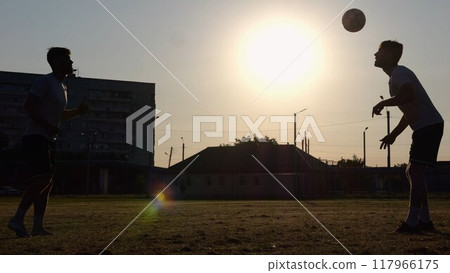 Silhouette of professional footballers kicking soccer ball with their heads on stadium at sunset. Two sportsmen showing tricks with ball while passing it to each other at field. Freestyle football 117966175
