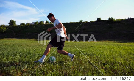 Professional footballer in uniform shows tricks with ball while running at green field. Sportsman kicks ball during jogging at meadow on sunny day. Young man training at stadium. Freestyle football Professional footballer in uniform shows tricks with ball while running at green field. Sportsman kicks ball during jogging at meadow on sunny day. Young man training at stadium. Freestyle football 117966247