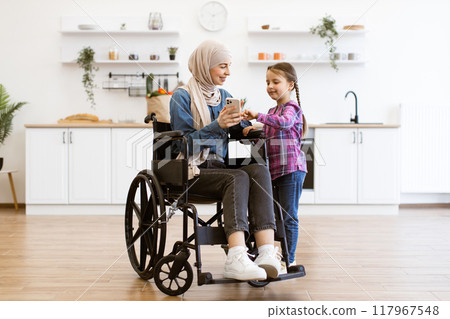 Muslim mother in wheelchair with daughter using mobile phone in kitchen 117967548