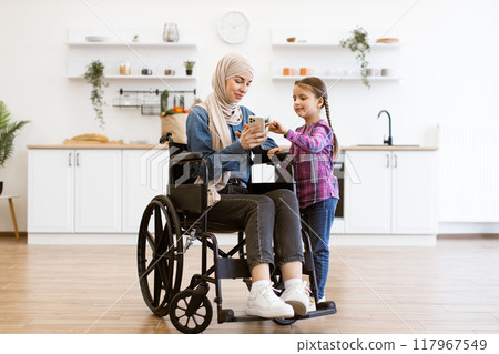 Muslim mother in wheelchair and daughter looking at smartphone in kitchen 117967549