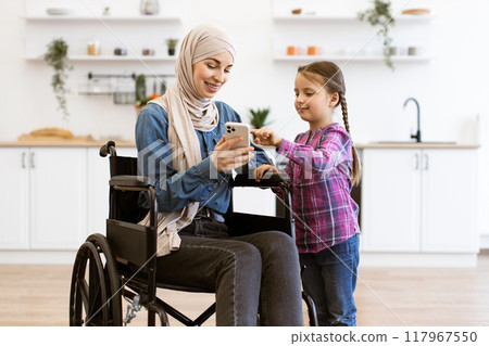 Muslim mother in wheelchair and daughter using mobile phone in kitchen 117967550