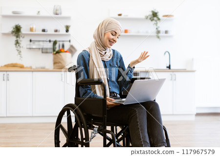 Smiling Muslim woman in wheelchair using laptop at home 117967579