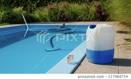 Pool cleaning hose and vacuum head with a large chemical container and water testing kit by the poolside, emphasizing maintenance and water quality. Pool cleaning hose and vacuum head with a large chemical container and water testing kit by the poolside, emphasizing maintenance and water quality. 117968754