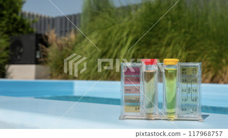 A close-up of a pool water testing kit displaying pH and chlorine levels, set on the edge of a pool with a blurred natural backdrop. 117968757